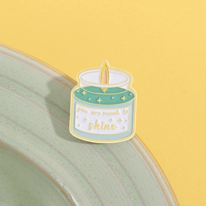 Enamel pin detail of a candle shape with text 'you are meant to shine' on yellow background