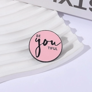 Broche ronde émaillée rose avec texte BE you TIFUL sur un support texturé blanc