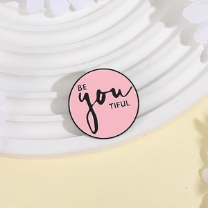 Broche ronde émaillée rose avec slogan BE YOU TIFUL sur support décoratif blanc