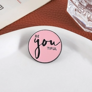 Broche ronde émaillée rose avec texte BE YOU TIFUL sur un plateau en céramique blanc