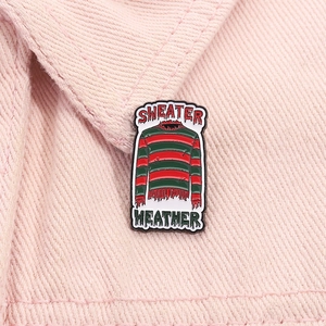 Enamel pin featuring a red and green striped sweater and Sweater Weather text on denim