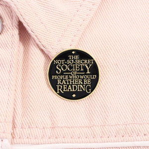 Detail of round black enamel book pin with gold inspirational quote on pink denim fabric