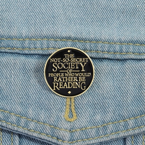 Round black enamel reading society brooch badge with gold lettering on blue denim fabric