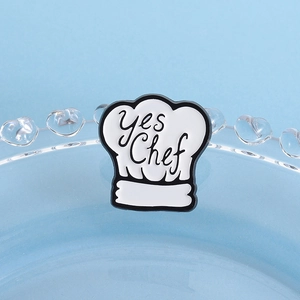 Detail shot of a white enamel chef hat pin featuring the black yes Chef text design
