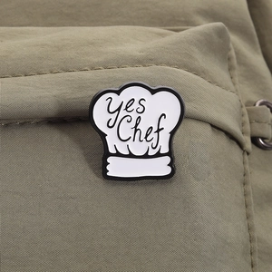 Detail of a white chef hat enamel pin with Yes Chef text on a green canvas backpack