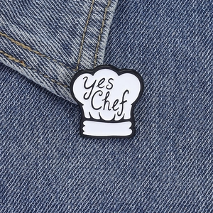 Detail of a white chef hat enamel pin with Yes Chef text on dark denim fabric