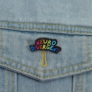 Spilla smaltata Neurodivergent arcobaleno su giacca di jeans con base nera e scritte colorate