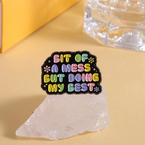 Detailed macro of enamel pin with quote 'Bit of a mess but doing my best' on white stone