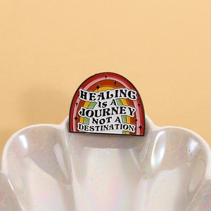 Macro detail of rainbow enamel pin with healing journey text on white shell tray