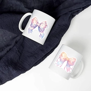 Top view of two white ceramic mugs with pastel watercolor bow designs on grey fabric