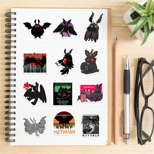 Flat lay of 12 Mothman graffiti stickers on a white notebook and wooden table