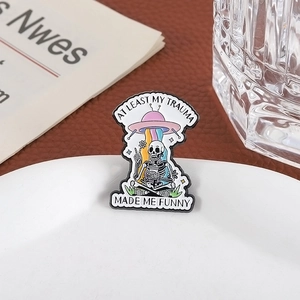 UFO enamel pin with skeleton and 'AT LEAST MY TRAUMA MADE ME FUNNY' quote on white plate