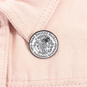 Detail of round enamel pin showing a skeleton reading a book on pink denim background