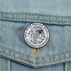 Skeleton reading book enamel pin with 'Read Banned Books' text on blue denim fabric