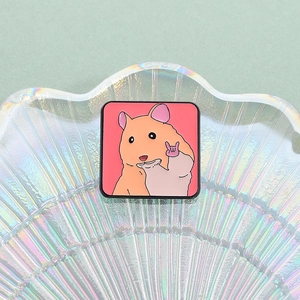 Broche en émail hamster mignon faisant un cœur posée sur un plateau en verre nacré