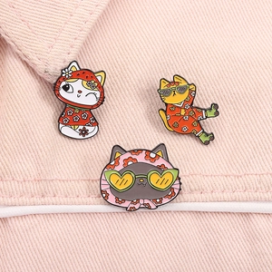 Three cute cartoon cat enamel pins displayed on pink denim fabric with vibrant colors