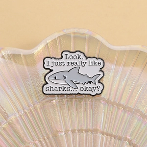 Detail shot of a cartoon shark enamel pin with quote displayed on a shell tray