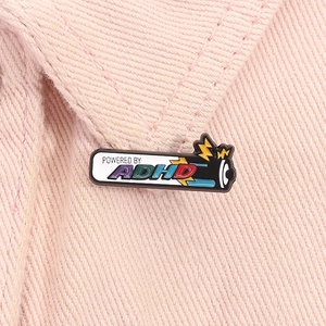 Detail of Powered by ADHD enamel pin with battery motif on pink fabric background