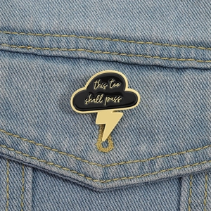 Black cloud and gold lightning enamel pin with 'this too shall pass' text on denim jacket