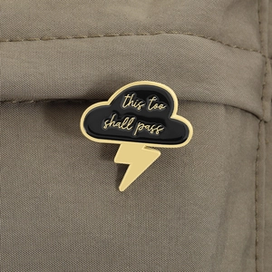 Detail view of a cloud and lightning enamel pin with 'this too shall pass' quote