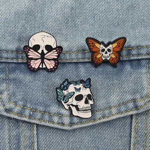 Three gothic skull butterfly enamel pins displayed on a blue denim jacket texture