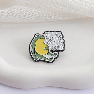 Funny green frog enamel pin with moody give me foody slogan on a white tray