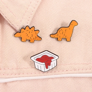 Detailed view of cartoon dinosaur chicken nugget and sauce enamel pins on pink fabric