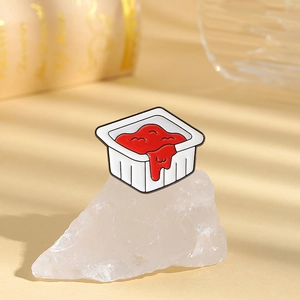 Macro view of a funny enamel pin shaped like a red sauce box on a white stone base