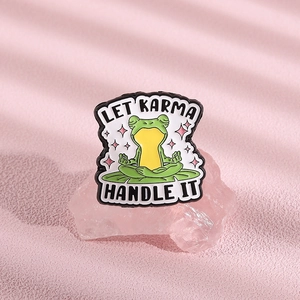 Macro shot of a green frog enamel pin on a pink crystal with Let Karma Handle It text