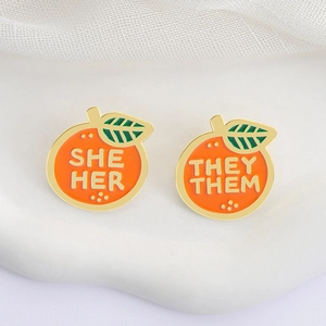 Orange enamel pins featuring SHE HER and THEY THEM pronouns on a wavy stand