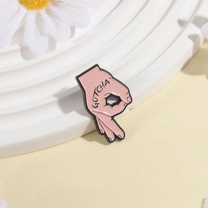 Makro-Detail einer rosa Emaille-Pin mit OK-Geste und GOTCHA-Schriftzug auf weißem Grund