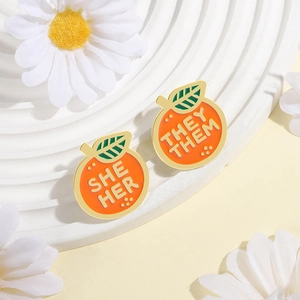 Close-up of orange enamel pins with SHE/HER and THEY/THEM pronouns and gold edges