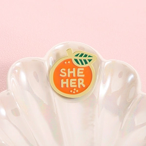 Detailed close-up of orange-shaped enamel pin featuring 'SHE HER' pronoun text