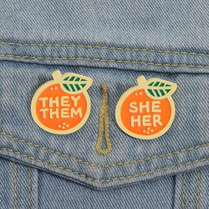 Detail of orange enamel pins with SHE HER and THEY THEM pronouns on denim jacket