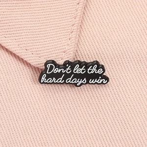 Detail of black enamel pin with Don't let the hard days win quote on pink fabric
