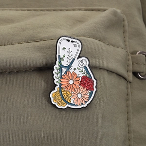 Detail einer kreativen Emaille-Brosche mit Stethoskop und Blumen an einer grünen Tasche