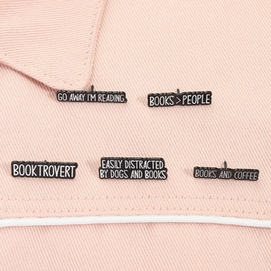 Detailed close-up of five black enamel book-themed pins on light pink fabric