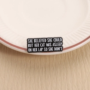 Detail of a black enamel brooch with cute cat-themed text on a white ceramic dish