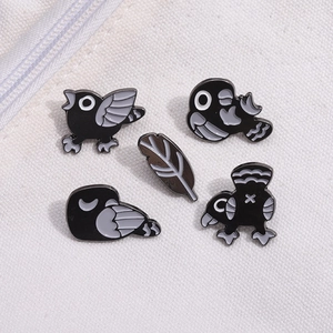 Close-up of five black cartoon bird enamel pins and a feather charm on white canvas