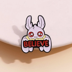 Cartoon white moth monster enamel pin with Believe banner on beige ceramic tray