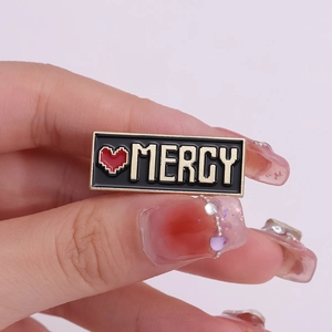 Spilla smaltata Mercy cuore pixel art con finiture dorate su fondo nero rettangolare