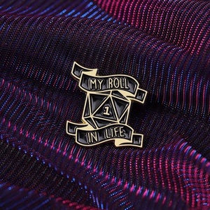Enamel pin with d20 dice and My Roll in Life text on dark metallic fabric background