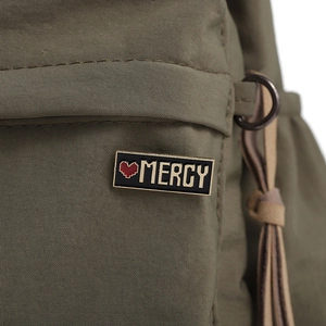 Dettaglio della spilla in smalto MERCY con cuore rosso su tessuto di borsa militare