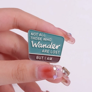 Broche en émail avec citation Wander Are Lost tenue sur un fond rose pastel