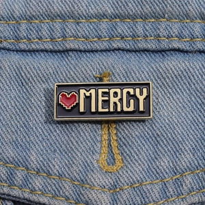 Dettaglio della spilla smaltata Mercy pixel art con cuore rosso su denim