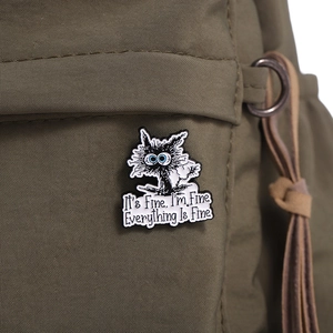 Macro view of black cat enamel pin with 'It's Fine' text on an olive canvas bag