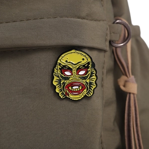 Detail of yellow creature mask enamel pin attached to a green canvas backpack pocket