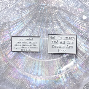 Set of two black and white enamel pins with humorous quotes in an iridescent shell dish