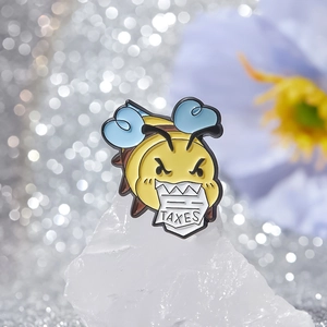 Cartoon bee enamel pin with blue wings biting a TAXES sign on white crystal stones