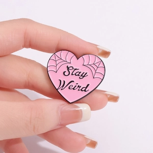 Pink heart enamel brooch with Stay Weird slogan and spider web design held in hand